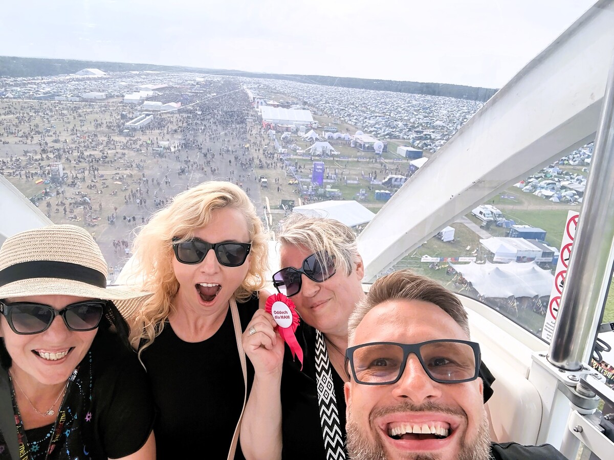 Cztery osoby robią selfie w gondoli diabelskiego młyna wysoko nad Pol’and’Rockiem. Za nimi rozciąga się ogromne pole namiotowe, a jedna z Mam trzyma przy twarzy różową przypinkę „Oddech dla MAM”.